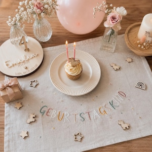 Könnte beinhalten: Ein beigefarbenes Leinen-Platzset mit der Stickerei GEBURTSTAGSKIND in Pastellfarben, einem Cupcake mit zwei brennenden Kerzen auf einem weißen Teller, einer Holzscheibe mit dem Namen Emilia und der Zahl 4, einem rosa Ballon und kleinen Holzfiguren.