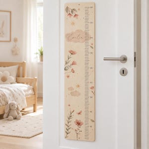 Puede incluir: Un medidor de altura vertical de madera montado en una puerta blanca, con una escala impresa de 80 a 160 centímetros. La superficie de madera clara está decorada con ilustraciones estilo acuarela de flores, soles, nubes y el nombre Emilia.