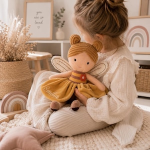 Könnte beinhalten: Eine Plüschpuppe mit Kordstruktur, bestehend aus einem senfgelben Kleid mit der Stickerei Emilia, einem braun-gelb gestreiften Oberteil, einer kleinen roten Blumenapplikation und weißen Flügeln. Die Puppe hat zwei kleine Haarknoten.