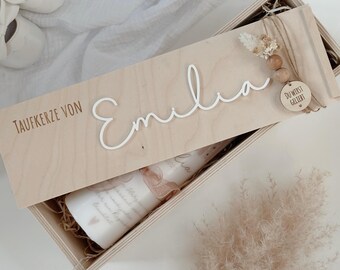 May include: A wooden box with a sliding lid, featuring the name "Emilia" in white script. The box contains a candle with a decorative ribbon and text. The lid also has the text "Taufkerze von" and a small wooden tag that says "Du wirst geliebt".