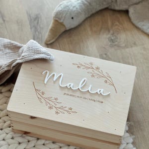 May include: A wooden keepsake box with a floral design and the name "Malia" engraved on the lid. The box also includes the date "27.03.2024", time "10:57 Uhr", weight "3,010g", and length "51cm".