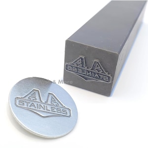 Könnte beinhalten: Ein Metallstempel mit dem Text "AA STAINLESS" in einem rechteckigen Rahmen, neben einer gestempelten Metallscheibe mit dem gleichen Design. Der Stempel ist grau und rechteckig, und die Scheibe ist silberfarben.