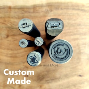 Op de afbeelding: Een set van zes op maat gemaakte metalen stempels met verschillende ontwerpen, waaronder een mes, een bierpul, een kroon, een logo en een beer. De stempels zijn in een cirkel op een houten oppervlak gerangschikt.