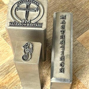 May include: Three rectangular metal stamps with raised designs. One features a cross and nails with the text HANDCRAFTED LEATHER WORKS LLC. Another shows a seahorse with a trident. The third is a long, narrow stamp with the word MASTERPIECE.