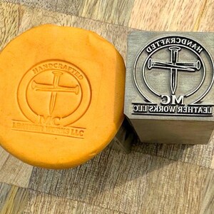 May include: A square metal stamp featuring a cross design made of two nails inside a circle with the text HANDCRAFTED MC LEATHER WORKS LLC, alongside a circular yellow clay impression showing the same embossed logo design in clear detail.
