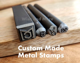 May include: Four metal stamps with different designs. The stamps are made of metal and have a silver finish. The designs include a geometric shape, a devil head, a stylized letter, and a symbol. The stamps are arranged in a row on a wooden surface. The text "Custom Made Metal Stamps" is printed below the stamps.