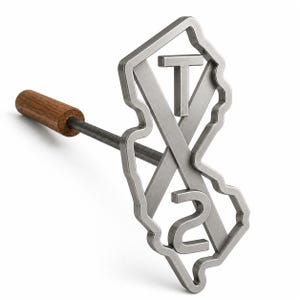 May include: A metal branding iron featuring a silver-toned outline of the state of Texas with the letters T and S and the number 2 integrated into the design. The tool has a long, thin metal rod attached to a cylindrical brown wooden handle.