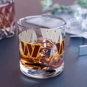 Könnte beinhalten: Ein klares Glas mit brauner Flüssigkeit und Eiswürfeln. Das Glas hat ein Design mit einem roten und gelben "W" mit einem Federmuster.