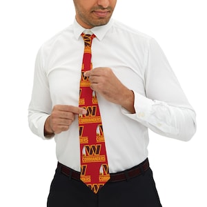 Op de afbeelding: Een rode stropdas met een herhalend patroon van het Washington Commanders-logo. Het logo heeft een bruine veer, een gouden 'W' en de woorden 'Commanders' in goud.
