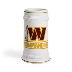 Puede incluir: Jarra de cerveza de cerámica blanca con borde dorado y un logotipo de los Washington Commanders en color burdeos y dorado. El logotipo presenta una "W" estilizada con un diseño de plumas.