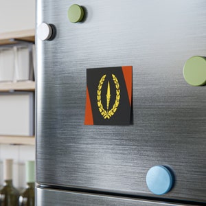 Puede incluir: Un imán cuadrado con fondo negro, que presenta un diseño de lanza dorada dentro de una corona de laurel. El diseño está flanqueado por rayas diagonales rojas. El imán está adherido a una puerta de nevera plateada con imanes de colores.