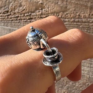 Op de afbeelding: Een zilverkleurige ring met een miniatuur theepot- en kopjesontwerp. De theepot is gedetailleerd met sierlijke patronen en verbonden met de kopjesring door een fijne ketting. De ring wordt op een vinger gedragen.