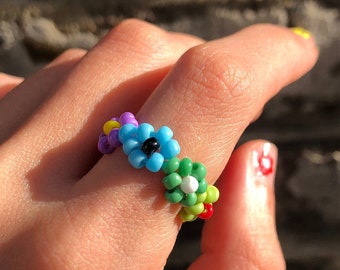 Op de afbeelding: Een kleurrijke kralenring met een bloemmotief. De ring is gemaakt van kleine, ronde kralen in tinten blauw, groen, geel en paars. Het midden van de bloem heeft een zwarte kraal.