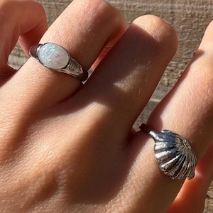 May include: Two silver rings on a hand. The ring on the index finger has a white oval stone. The ring on the middle finger is shaped like a seashell.