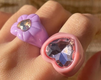 May include: Two rings with large, clear rhinestones. The ring on the left is purple and shaped like a flower. The ring on the right is pink and shaped like a heart.