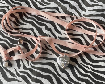 Op de afbeelding: Een roze lint choker ketting met een zilveren hartvormig medaillon en een ster bedel. Het medaillon heeft gegraveerde tekst. Een kleine parel bedel hangt aan het lint. De ketting wordt gepresenteerd op een zwart-wit zebra gestreepte achtergrond.