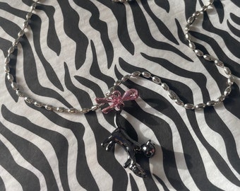 May include: A silver chain necklace with a black cat charm and a pink bow. The necklace is on a zebra print background.