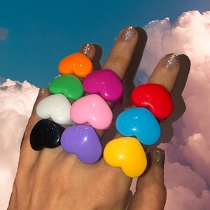 May include: A set of ten heart-shaped rings in various colours, including pink, red, yellow, green, blue, purple, orange, white, and black. The rings are stacked on a hand, with the tips of the fingers visible.