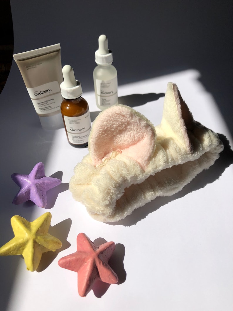 Puede incluir: Una diadema blanca con orejeras rosas, tres bombas de ba&ntilde;o en forma de estrella en morado, amarillo y rosa, y tres botellas de productos para el cuidado de la piel de The Ordinary.