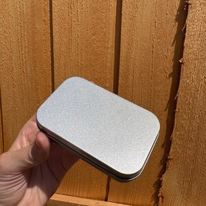 Puede incluir: Una caja de metal rectangular con esquinas redondeadas y un acabado liso de color plateado. El recipiente cuenta con una tapa plana que encaja firmemente sobre la base, presentando una superficie met&aacute;lica limpia con una textura sutil.