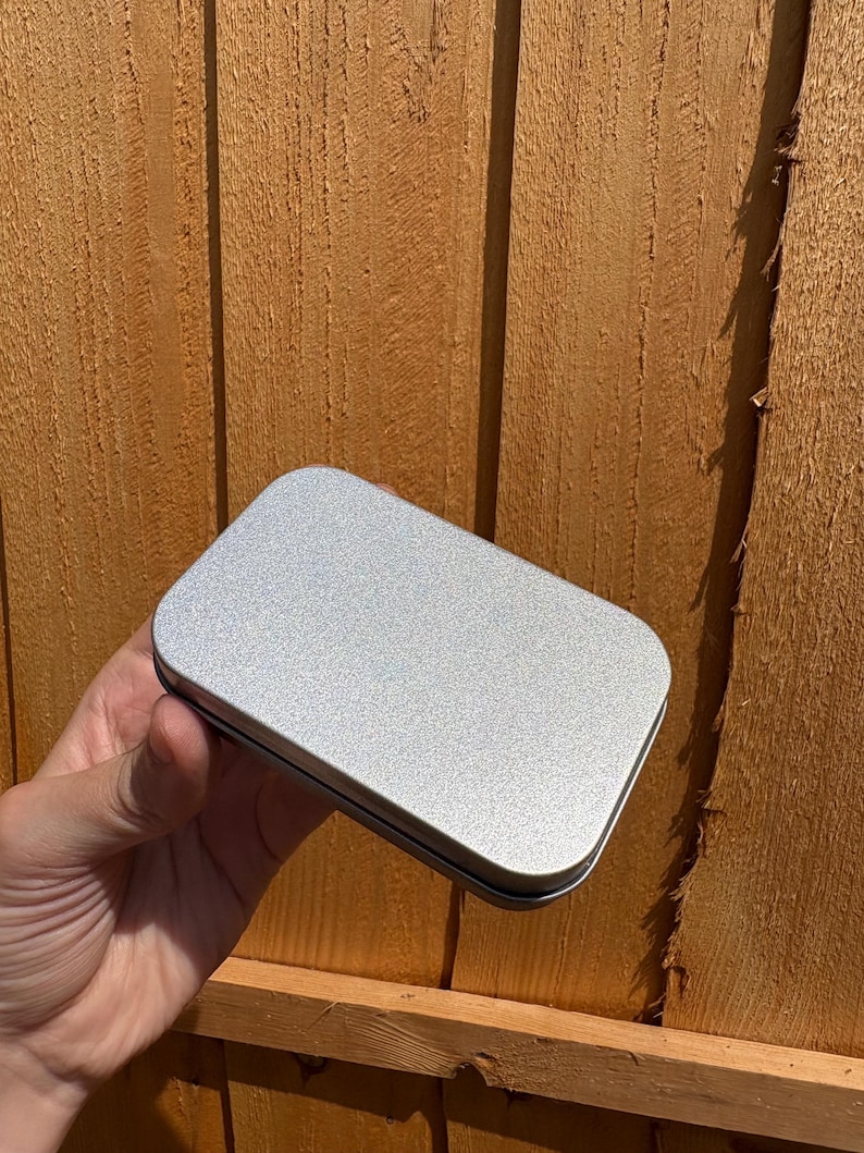 Puede incluir: Una caja de metal rectangular con esquinas redondeadas y un acabado liso de color plateado. El recipiente cuenta con una tapa plana que encaja firmemente sobre la base, presentando una superficie met&aacute;lica limpia con una textura sutil.
