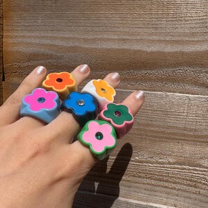 Op de afbeelding: Een hand toont zeven dikke bloemvormige ringen in verschillende kleuren: oranje, geel, blauw, wit, roze, groen en lichtblauw. Elke ring heeft een kleine, sprankelende edelsteen in het midden. De ringen zijn gemaakt van glanzend materiaal.