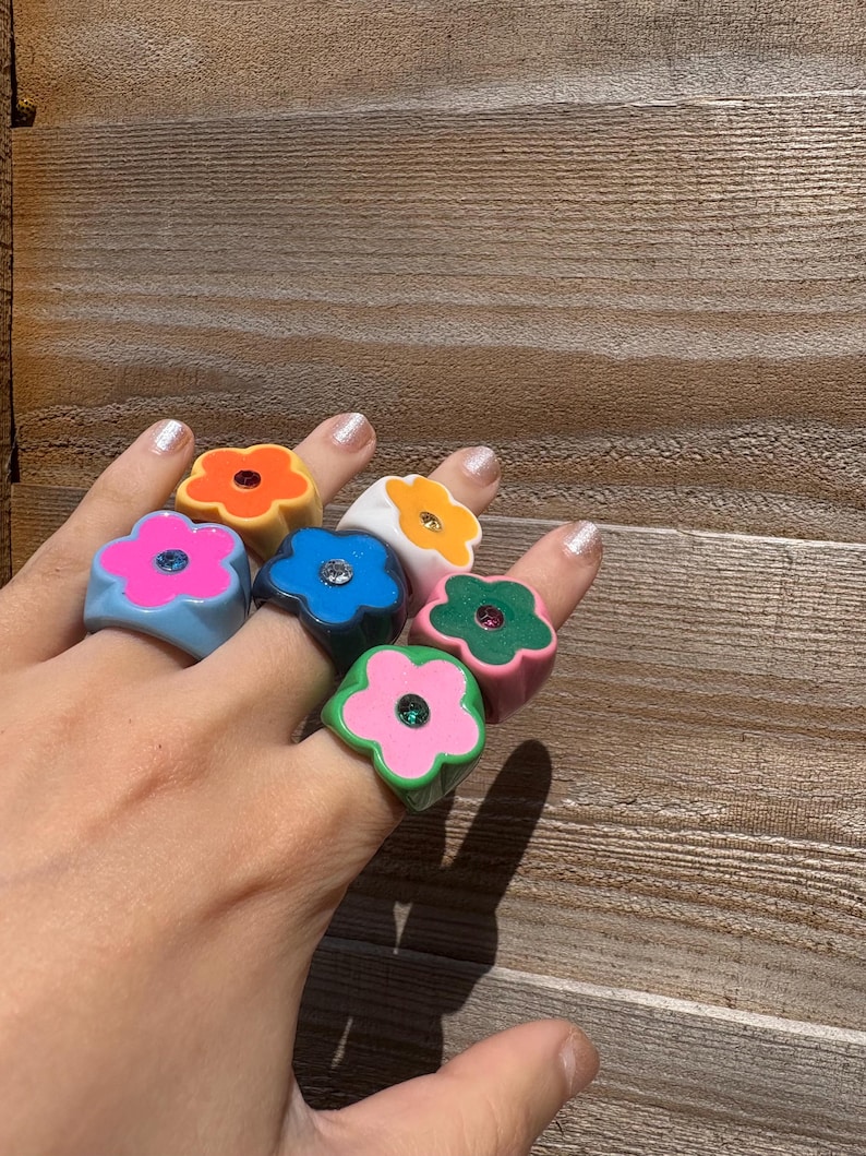 Op de afbeelding: Een hand toont zeven dikke bloemvormige ringen in verschillende kleuren: oranje, geel, blauw, wit, roze, groen en lichtblauw. Elke ring heeft een kleine, sprankelende edelsteen in het midden. De ringen zijn gemaakt van glanzend materiaal.