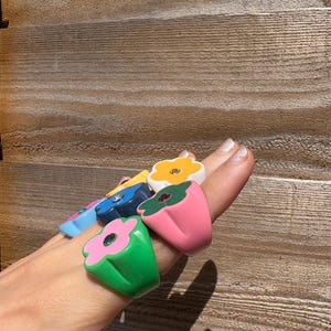 Op de afbeelding: Een verzameling kleurrijke bloemvormige ringen. De ringen zijn in verschillende kleuren, waaronder groen, roze, blauw en geel, met een contrasterende kleur voor de bloemblaadjes. Elke ring heeft een edelsteen in het midden.