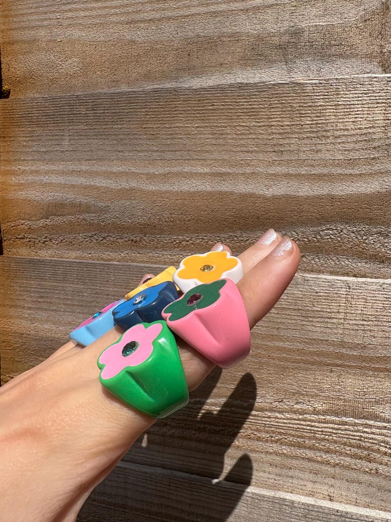 Op de afbeelding: Een verzameling kleurrijke bloemvormige ringen. De ringen zijn in verschillende kleuren, waaronder groen, roze, blauw en geel, met een contrasterende kleur voor de bloemblaadjes. Elke ring heeft een edelsteen in het midden.