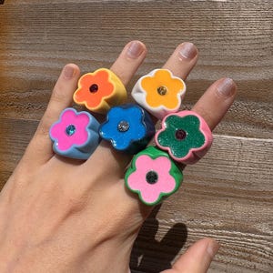 Op de afbeelding: Een hand die zeven bloemvormige ringen in verschillende kleuren toont. De ringen zijn geel, wit, blauw, groen en roze, elk met een contrasterende middensteen. De ringen zijn gemaakt van een glanzend materiaal.