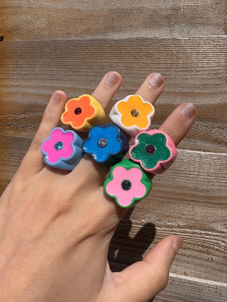 Op de afbeelding: Een hand die zeven bloemvormige ringen in verschillende kleuren toont. De ringen zijn geel, wit, blauw, groen en roze, elk met een contrasterende middensteen. De ringen zijn gemaakt van een glanzend materiaal.