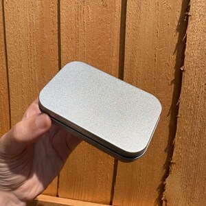 Puede incluir: Una caja met&aacute;lica rectangular de color plateado con esquinas redondeadas y un acabado mate liso. El recipiente cuenta con una tapa plana sencilla que encaja firmemente sobre la base, presentando una superficie met&aacute;lica limpia y uniforme.