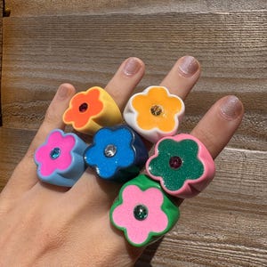 Op de afbeelding: Een hand toont zes kleurrijke bloemvormige ringen. De ringen zijn in tinten roze, blauw, groen, geel en wit, elk met een contrasterende middensteen. De ringen zijn gemaakt van een glanzend materiaal.