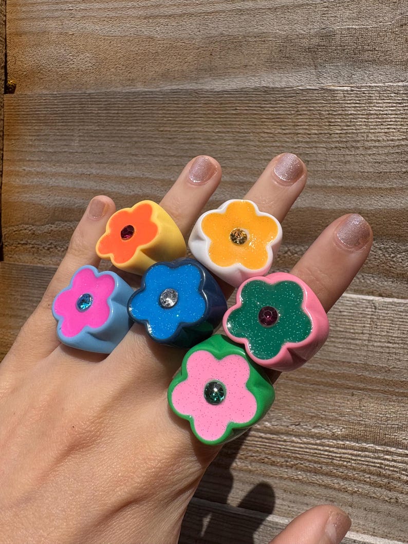 Op de afbeelding: Een hand toont zes kleurrijke bloemvormige ringen. De ringen zijn in tinten roze, blauw, groen, geel en wit, elk met een contrasterende middensteen. De ringen zijn gemaakt van een glanzend materiaal.