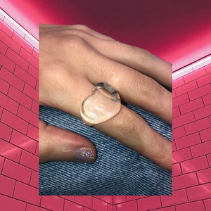 Puede incluir: Un anillo transparente en forma de corazón con un acabado ligeramente esmerilado. El anillo se lleva en un dedo con un esmalte de uñas brillante.