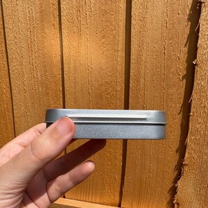 Puede incluir: Una caja rectangular de metal plateado con un acabado liso y una tapa ajustada. El recipiente presenta esquinas redondeadas y un perfil delgado, mostrando un brillo met&aacute;lico que refleja la luz en su superficie. El objeto se presenta con un dise&ntilde;o sencillo y funcional.