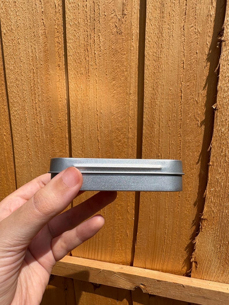 Puede incluir: Una caja rectangular de metal plateado con un acabado liso y una tapa ajustada. El recipiente presenta esquinas redondeadas y un perfil delgado, mostrando un brillo met&aacute;lico que refleja la luz en su superficie. El objeto se presenta con un dise&ntilde;o sencillo y funcional.