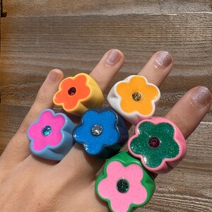 Op de afbeelding: Een hand toont zes bloemvormige ringen in verschillende kleuren. De ringen zijn geel, oranje, wit, blauw, roze en groen. Elke ring heeft een edelsteen in het midden. De ringen zijn gemaakt van glanzend materiaal.