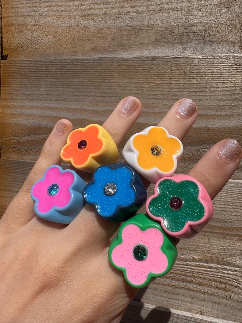 Op de afbeelding: Een hand toont zes bloemvormige ringen in verschillende kleuren. De ringen zijn geel, oranje, wit, blauw, roze en groen. Elke ring heeft een edelsteen in het midden. De ringen zijn gemaakt van glanzend materiaal.