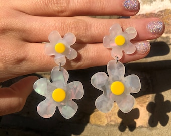 Op de afbeelding: Een paar bloemvormige oorbellen. De oorbellen hebben een doorschijnend, iriserend wit bloemblaadjesontwerp met een heldergele kern. De oorbellen zijn opgehangen aan een kleine zilveren haak.