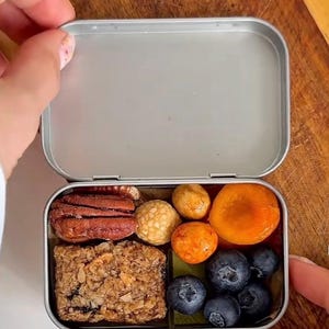 Op de afbeelding: Een rechthoekige zilveren metalen blikken doos gevuld met diverse snacks, waaronder een textuurrijke mueslireep, bruine pecannoten, ronde beige en oranje hapjes, een gedroogde abrikoos en verschillende verse donkerblauwe bosbessen.