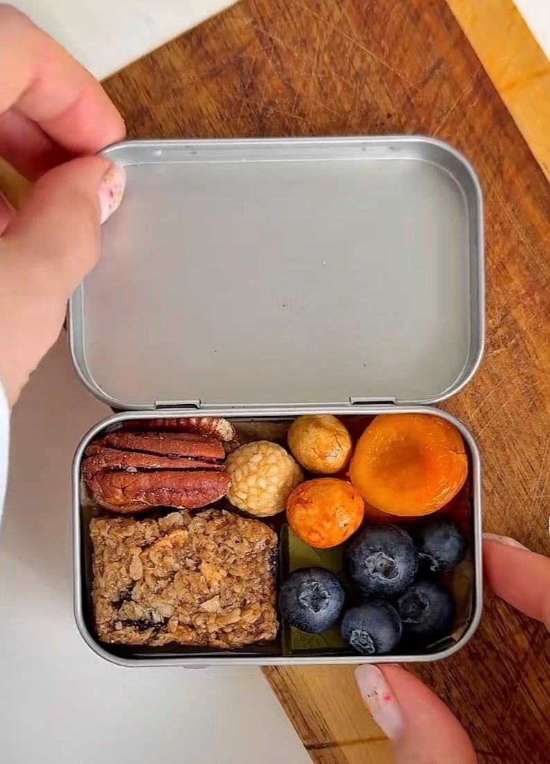 Puede incluir: Un recipiente rectangular de metal plateado lleno de una variedad de bocadillos que incluyen una barra de granola texturizada, nueces pecanas marrones, bocados redondos beige y naranjas, un albaricoque seco y varios ar&aacute;ndanos frescos azul oscuro.
