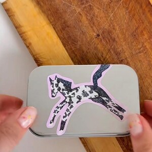 Puede incluir: Una caja rectangular de metal plateado con una pegatina de un caballo saltando con pelaje moteado en blanco y negro. La ilustraci&oacute;n del caballo tiene un contorno rosa claro y est&aacute; colocada en el centro de la tapa lisa y reflectante.