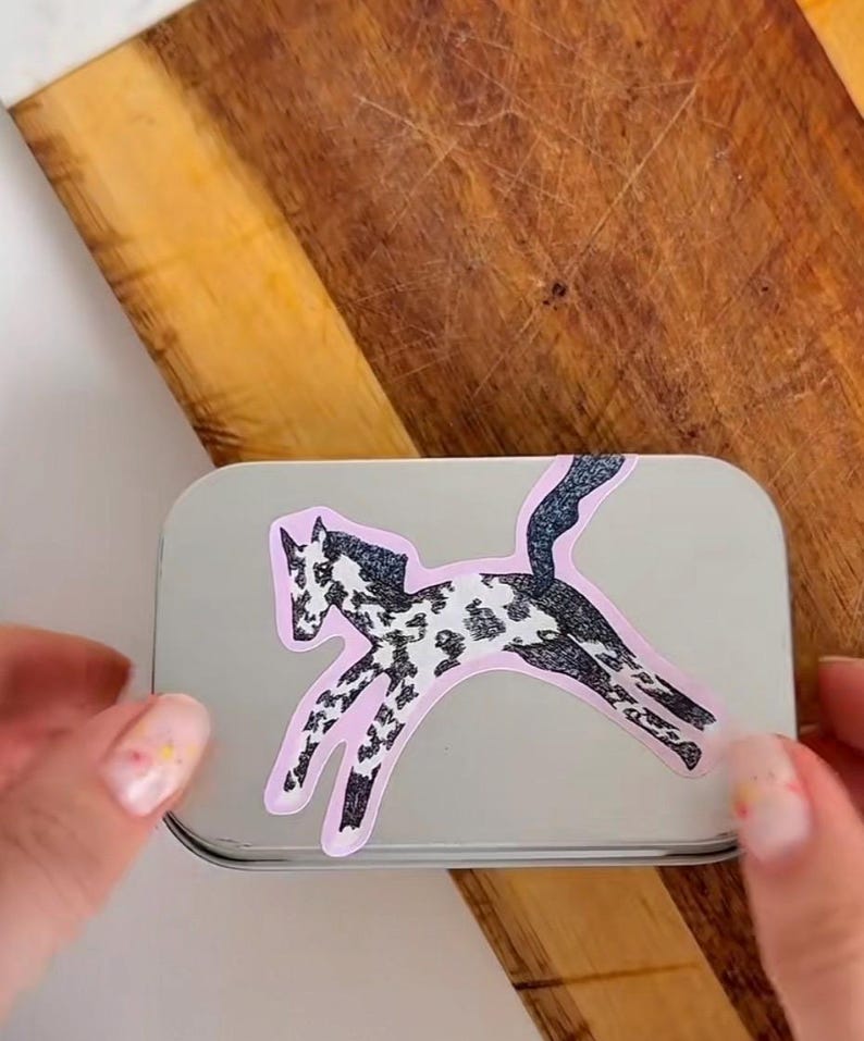 Puede incluir: Una caja rectangular de metal plateado con una pegatina de un caballo saltando con pelaje moteado en blanco y negro. La ilustraci&oacute;n del caballo tiene un contorno rosa claro y est&aacute; colocada en el centro de la tapa lisa y reflectante.