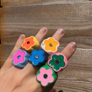 Op de afbeelding: Een hand toont zeven bloemvormige ringen in verschillende kleuren. De ringen zijn oranje, geel, blauw, roze en groen. Elke ring heeft een kleine, sprankelende edelsteen in het midden. De ringen zijn gemaakt van glanzend materiaal.