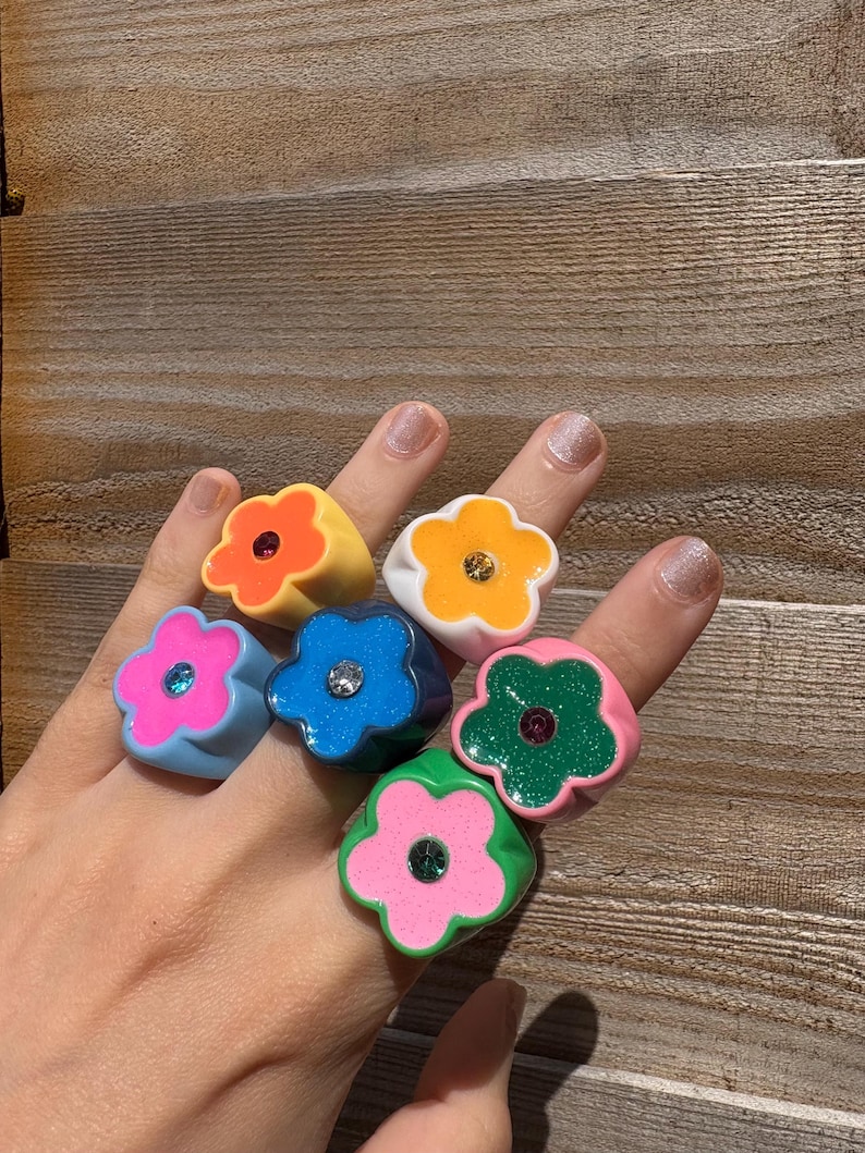 Op de afbeelding: Een hand toont zeven bloemvormige ringen in verschillende kleuren. De ringen zijn oranje, geel, blauw, roze en groen. Elke ring heeft een kleine, sprankelende edelsteen in het midden. De ringen zijn gemaakt van glanzend materiaal.