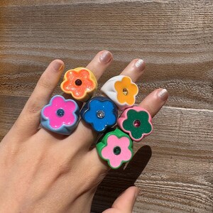 Op de afbeelding: Een hand toont zeven bloemvormige ringen in levendige kleuren. De ringen zijn oranje, wit, roze, blauw en groen. Elke ring heeft een kleine edelsteen in het midden. De ringen zijn gemaakt van glanzend materiaal.