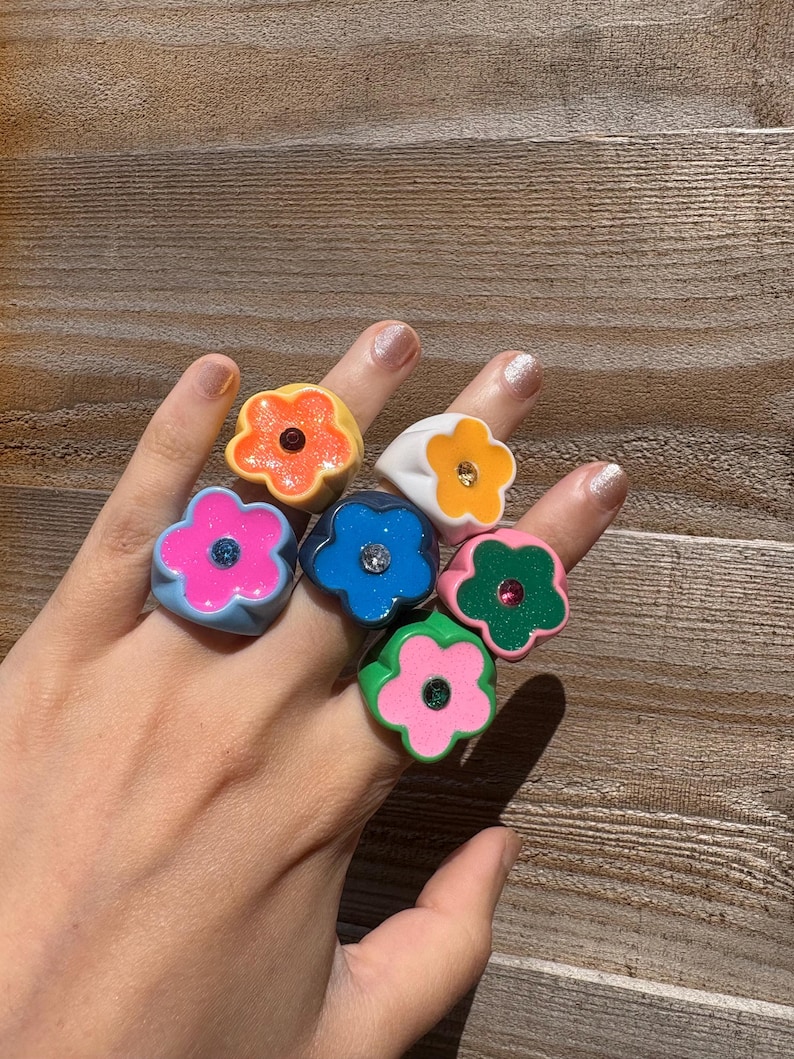 Op de afbeelding: Een hand toont zeven bloemvormige ringen in levendige kleuren. De ringen zijn oranje, wit, roze, blauw en groen. Elke ring heeft een kleine edelsteen in het midden. De ringen zijn gemaakt van glanzend materiaal.