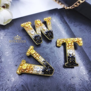 Peut inclure: Gros plan sur quatre épingles à lettres perlées : deux W, un T et un V. Les lettres sont dorées, noires et en cristal transparent. Les épingles sont posées sur une surface gris foncé avec un motif floral. La marque "NYNAZZEN" est visible.