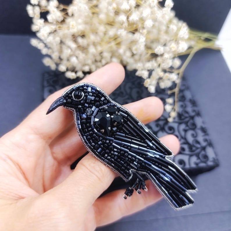 May include: A black beaded crow brooch with a black crystal eye and a black crystal heart on its chest.